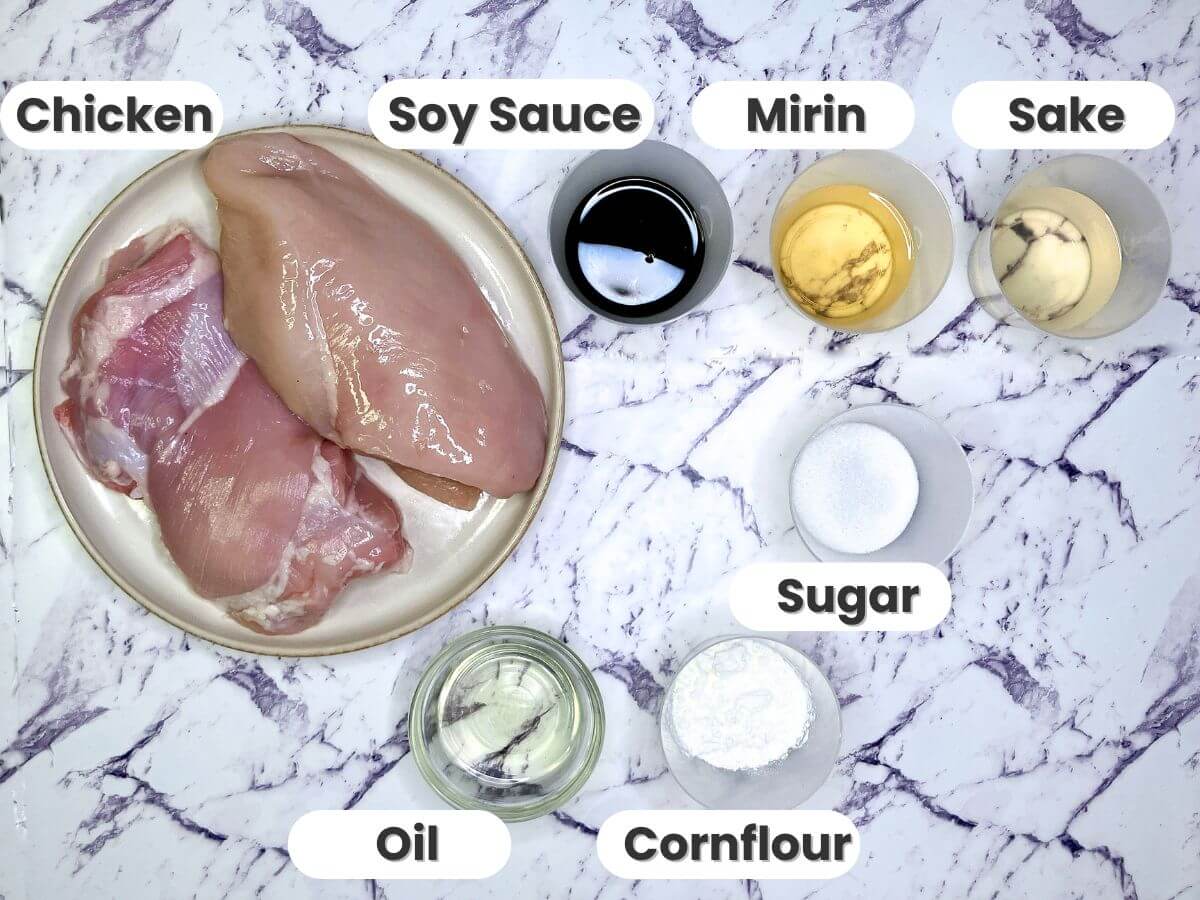"Raw chicken breast and thigh on a plate surrounded by labeled ingredients for teriyaki chicken: soy sauce, mirin, sake, sugar, oil, and cornflour on a marble background."