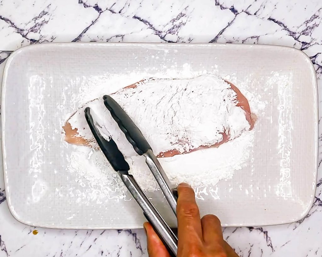 "Dredging chicken breast in cornflour using tongs on white plate."