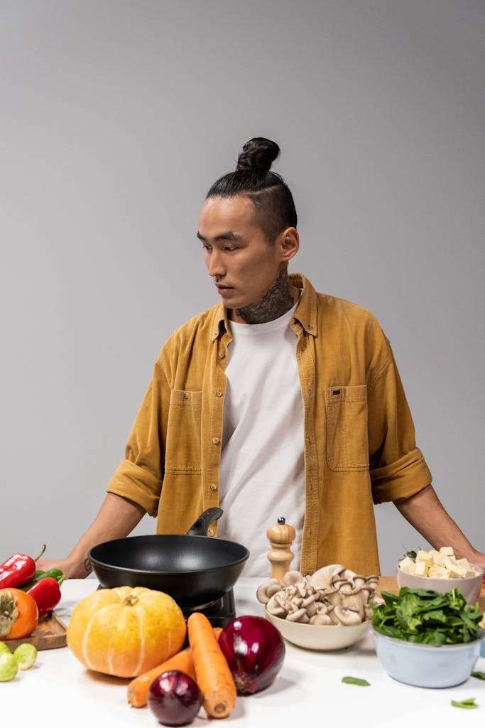 Tattooed chef with man bun cooking with fresh vegetables and mushrooms indoors.