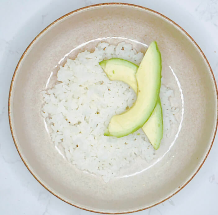 Sliced avocado placed on a bed of steamed rice as part of salmon bowl assembly.