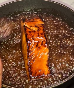 Basting salmon with thickened ginger teriyaki sauce while it cooks in a pan.