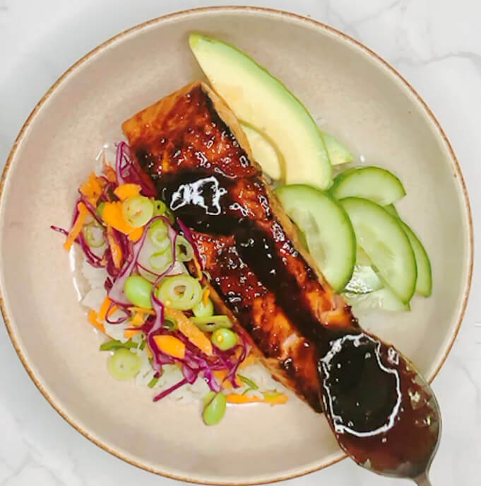 Drizzling thick teriyaki sauce over glazed salmon in a finished rice bowl.