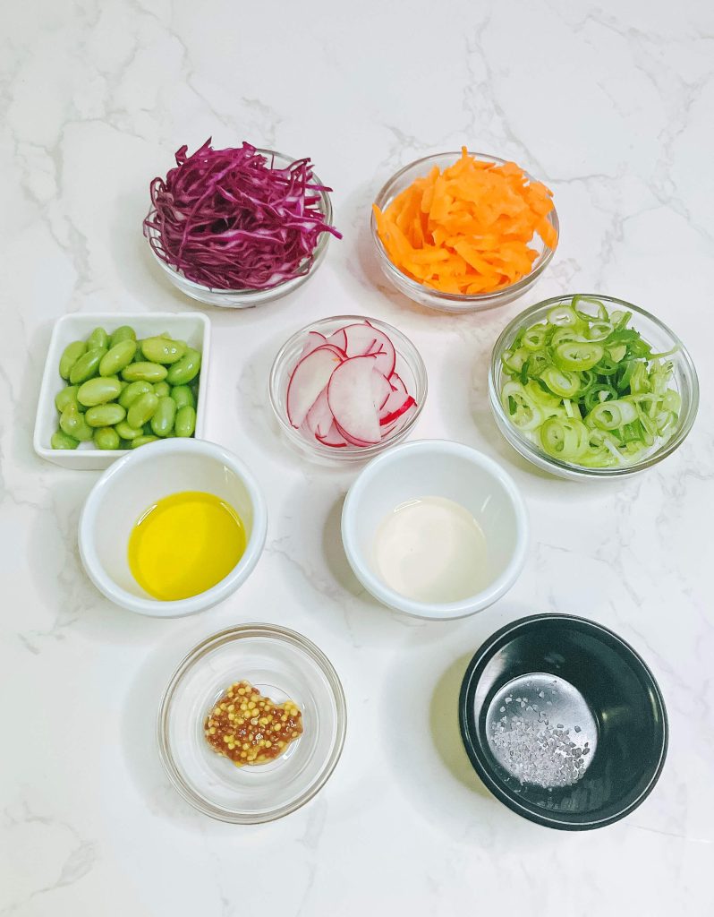 Flat lay of fresh salad ingredients for a salmon bowl, including cabbage, radish, edamame, carrot, and dressing.