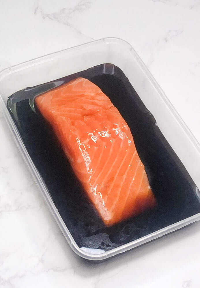 Salmon fillet marinating in pantry teriyaki sauce inside a plastic container.