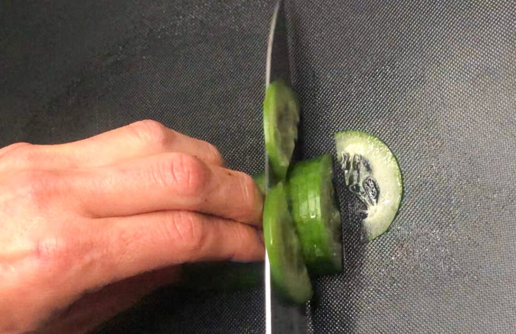 Slicing fresh cucumber with a knife for topping a salmon rice bowl.