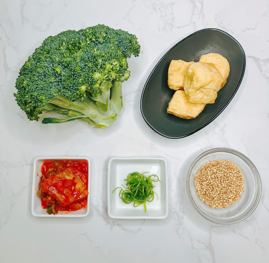 Flatlay of teriyaki broccoli tofu bowl ingredients including broccoli, tofu, kimchi, nori, sesame, and pantry sauce