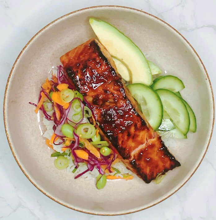 Teriyaki-glazed salmon served over rice with avocado, cucumber, and colorful salad.
