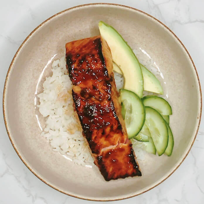 Glazed teriyaki salmon fillet placed on top of rice with avocado and cucumber.