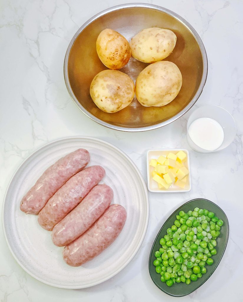 Flatlay of ingredients for bangers and mash, including raw sausages, unpeeled potatoes, butter, green peas, and onion on a white marble background.