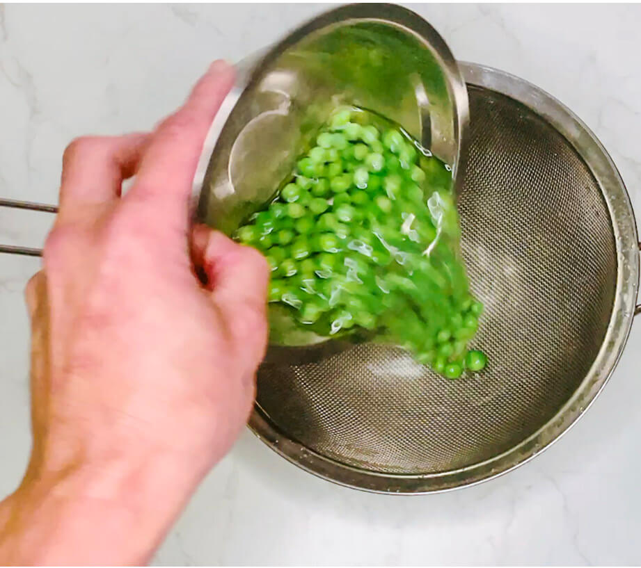 Hand using a fine mesh strainer to drain warmed frozen peas after heating in hot water.