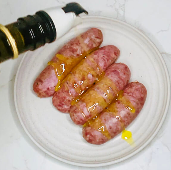 Pouring oil over raw sausages arranged on a plate to prepare for air frying in the bangers and mash recipe.