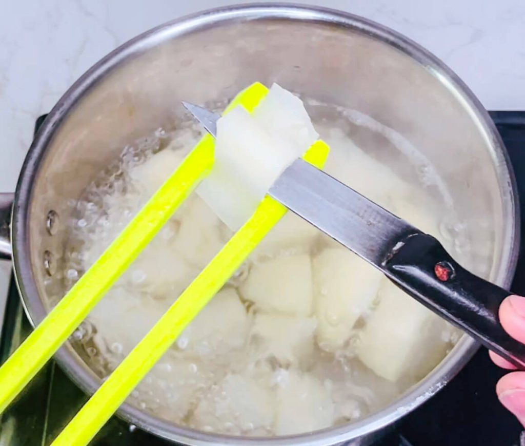 Hand checking doneness of boiling potatoes with knife to see if they’re soft and ready for mashing.