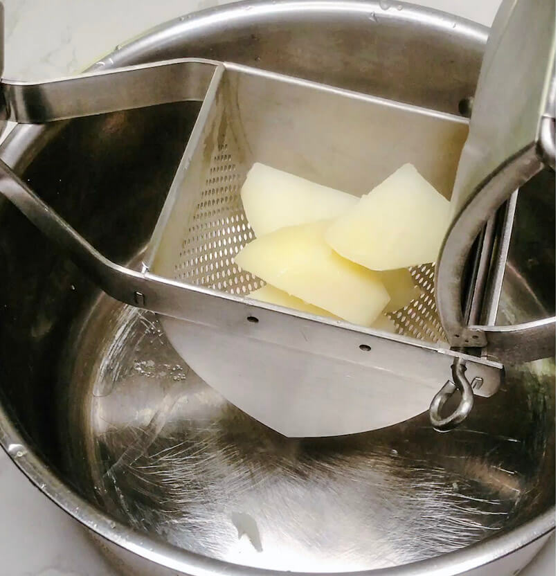 Pressing boiled potatoes through a metal potato ricer with butter into a saucepan to prepare mashed potatoes.