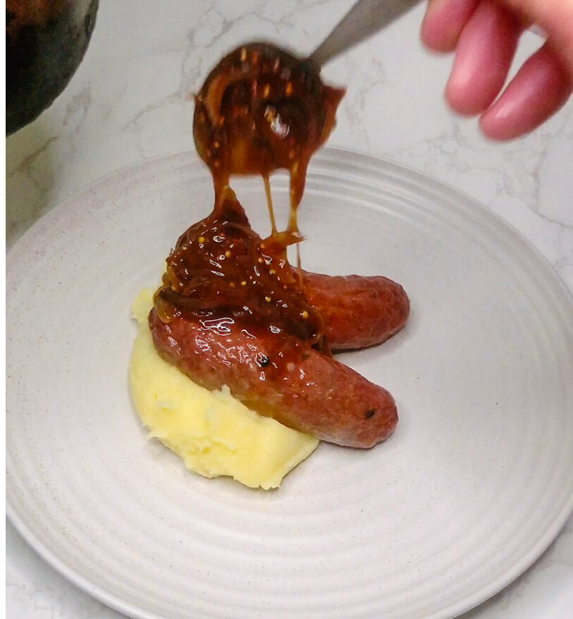 Pouring teriyaki onion gravy over sausages and mash for a savory finishing touch.
