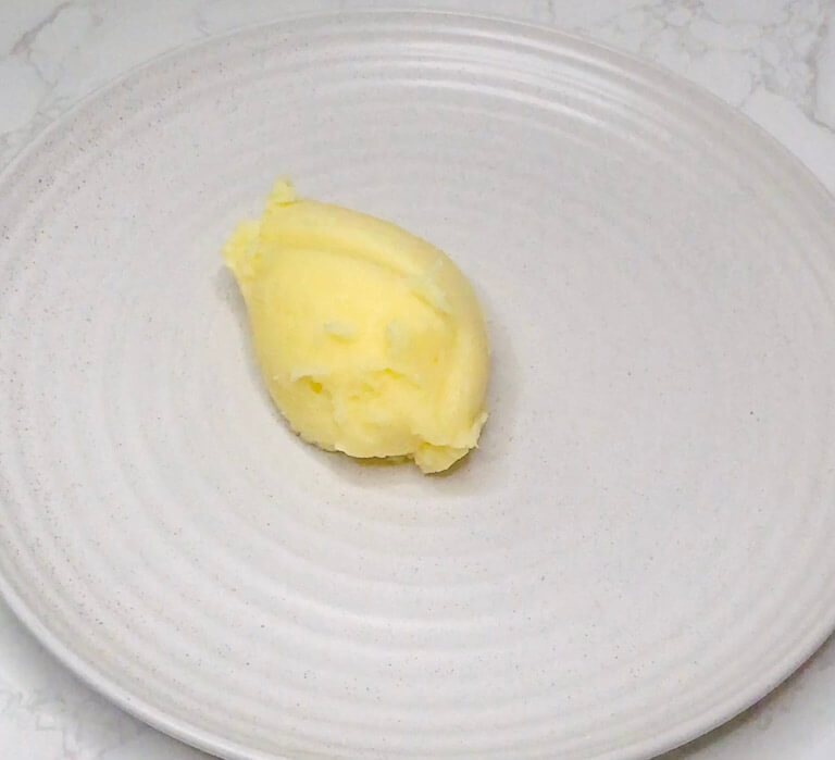 A single scoop of finished mashed potatoes plated in the center of a round ceramic dish, ready to be topped with sausages and gravy.