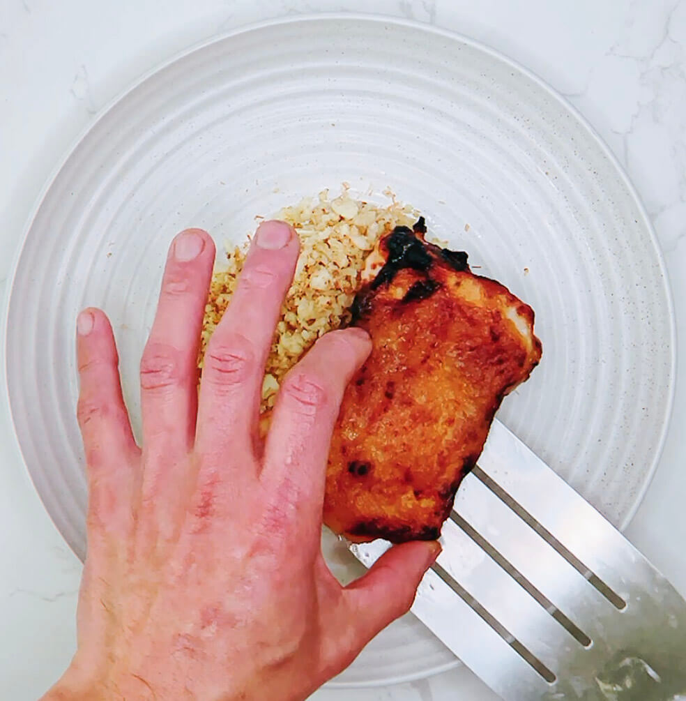 Hand placing a piece of miso chicken on top of cauliflower rice in a serving bowl.