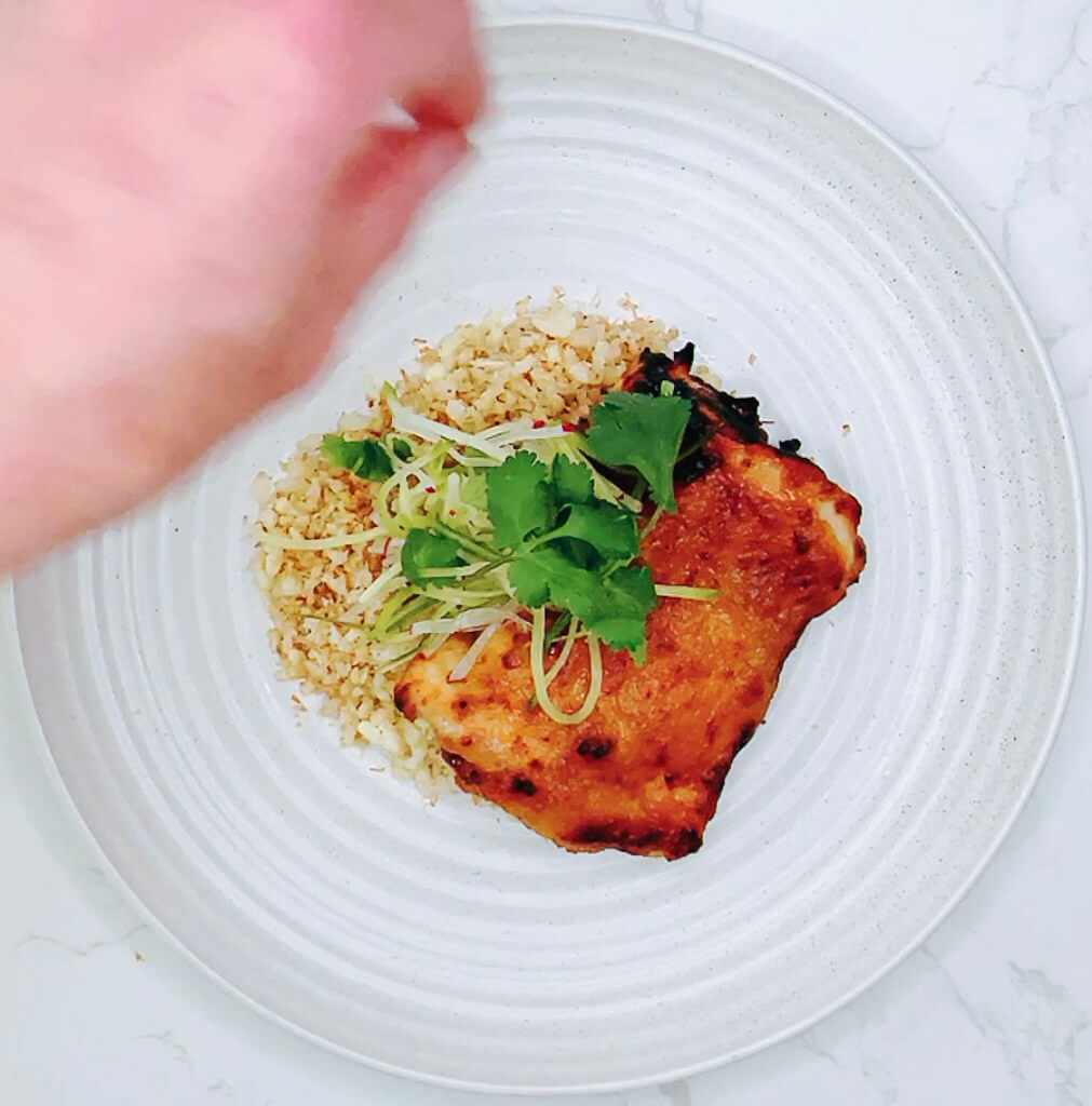 : Hand placing salad garnish beside miso chicken and cauliflower rice in a serving bowl.