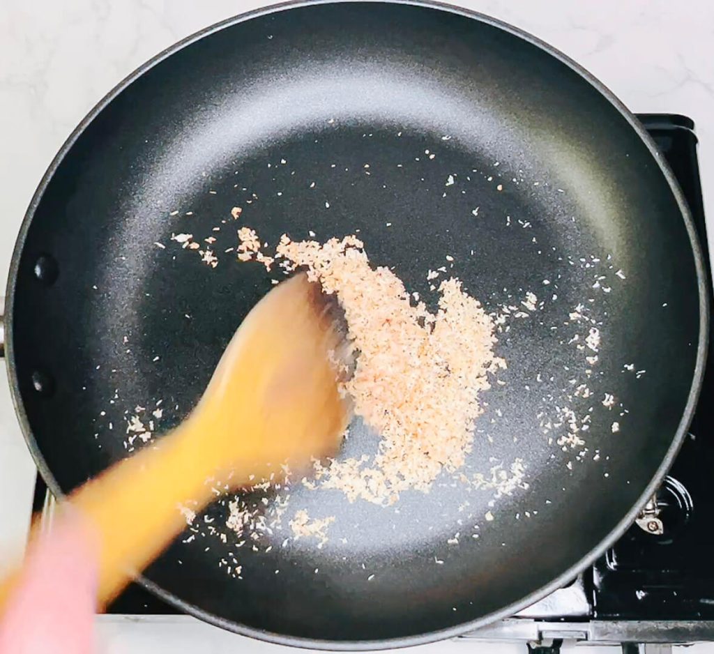 Wooden spoon stirring coconut in pan as it turns golden.