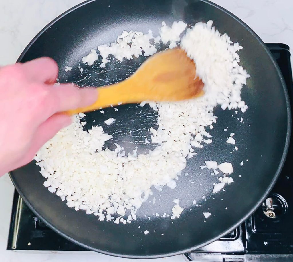 Riced cauliflower being stirred into coconut milk in the pan.