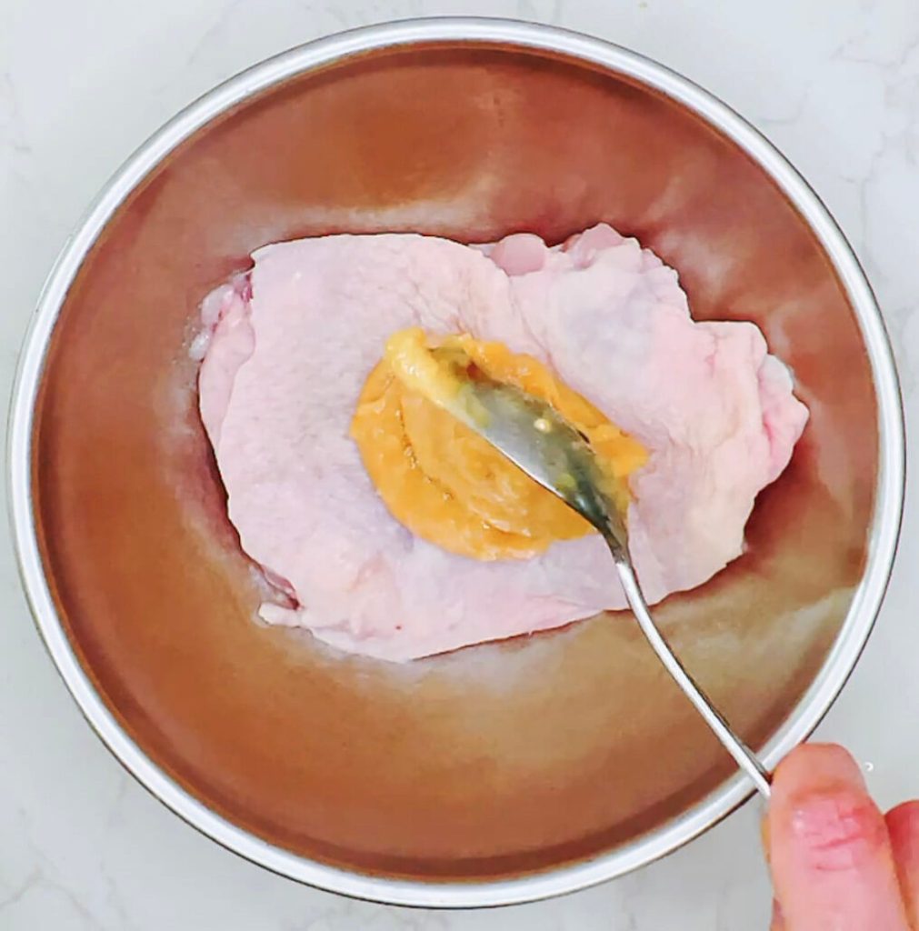 Pouring miso marinade over raw chicken thigh in a bowl.