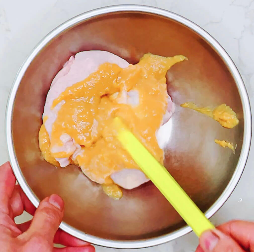 Spreading miso marinade over raw chicken thigh with a spoon in a bowl.