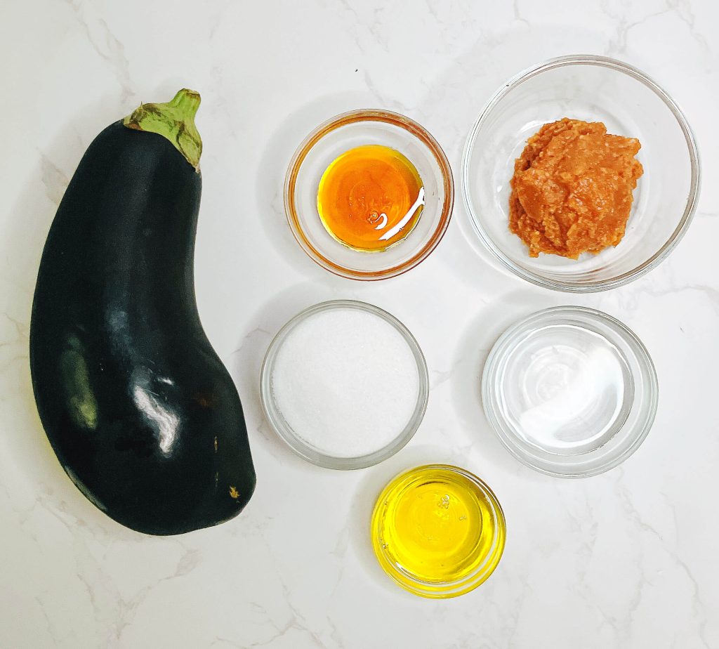 Top-down view of miso eggplant ingredients including eggplant, miso paste, golden syrup, sugar, water, and oil on a white background