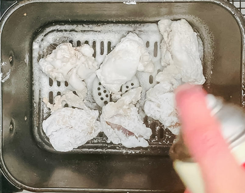Hand spraying oil over coated karaage chicken pieces in the air fryer basket."