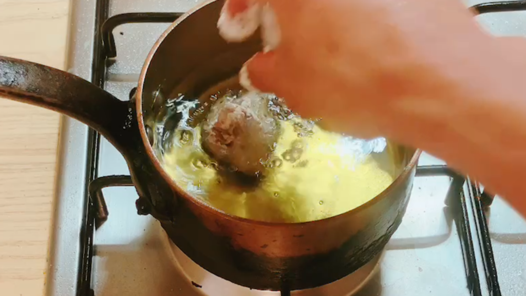 Hand lowering marinated pork pieces into hot oil for deep-frying, showing the start of the frying process for sweet and sour pork (Subuta).