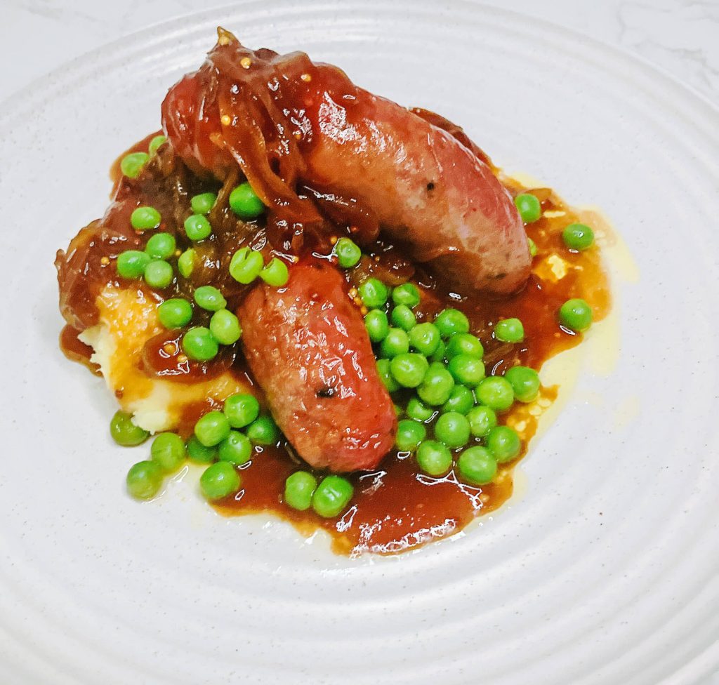Bangers and mash with teriyaki onion gravy, served in a ceramic bowl with mashed potatoes, caramelized onions, and peas — a Japanese-inspired comfort food twist.